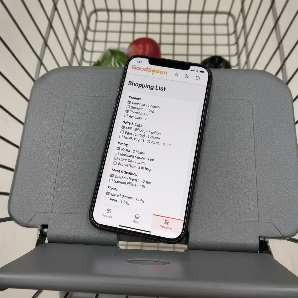 Smartphone with GoodSpoon shopping list displayed, sitting in grocery cart showing organized ingredients by category